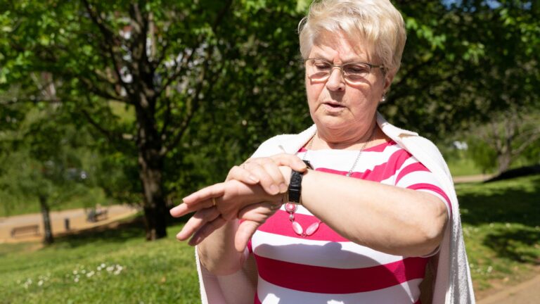 Aeltere Frau mit Brille schaut im Park auf ihre Armbanduhr