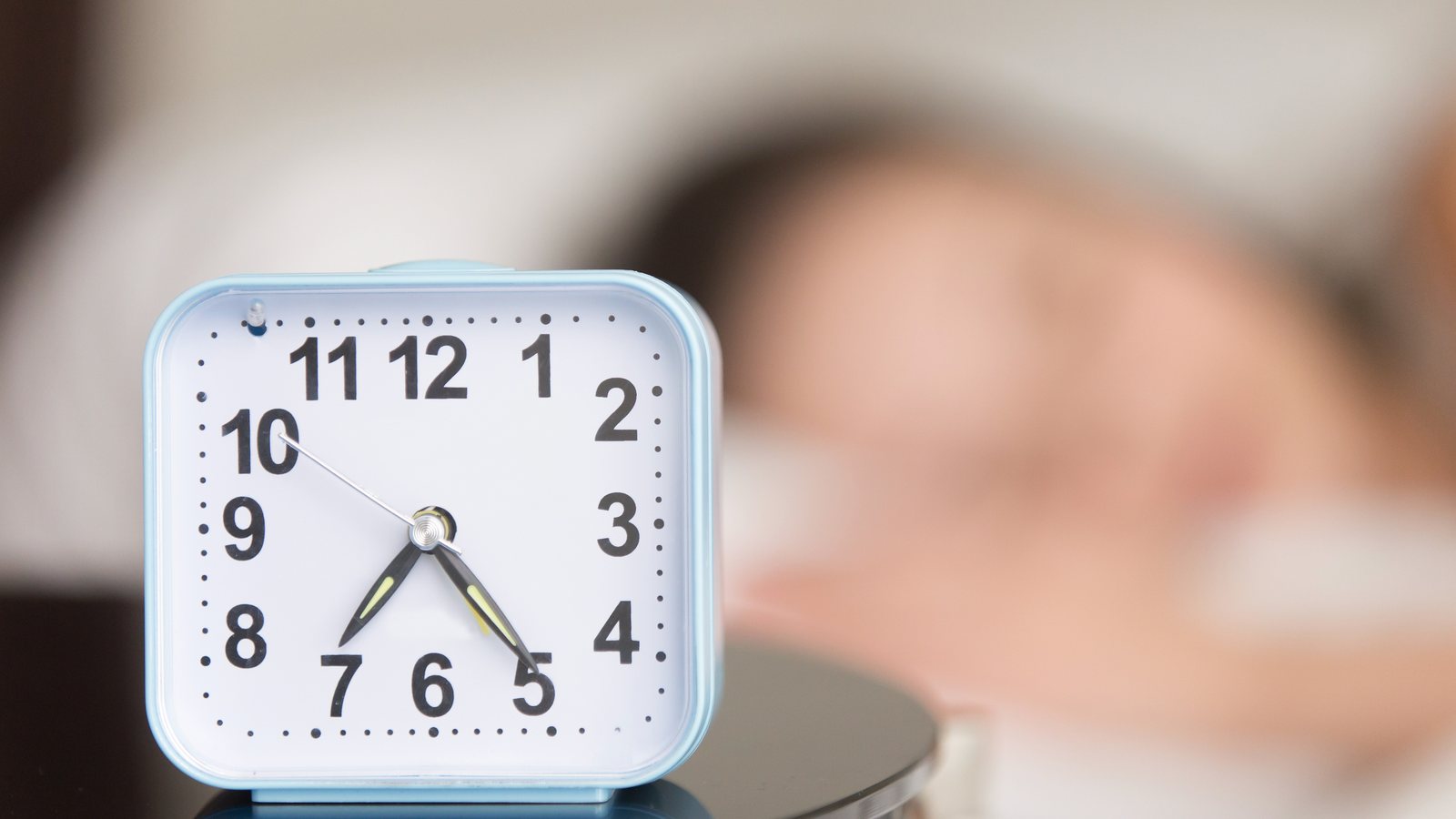 Weisser Wecker mit grossen, gut lesbaren Ziffern steht auf dem Nachttisch, im Hintergrund schlaeft eine Person