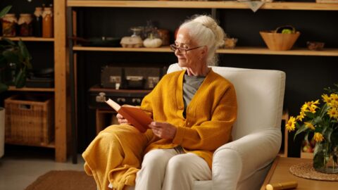 Aeltere Frau mit Brille und gelber Strickjacke sitzt mit einem Buch in einem hellen Sessel