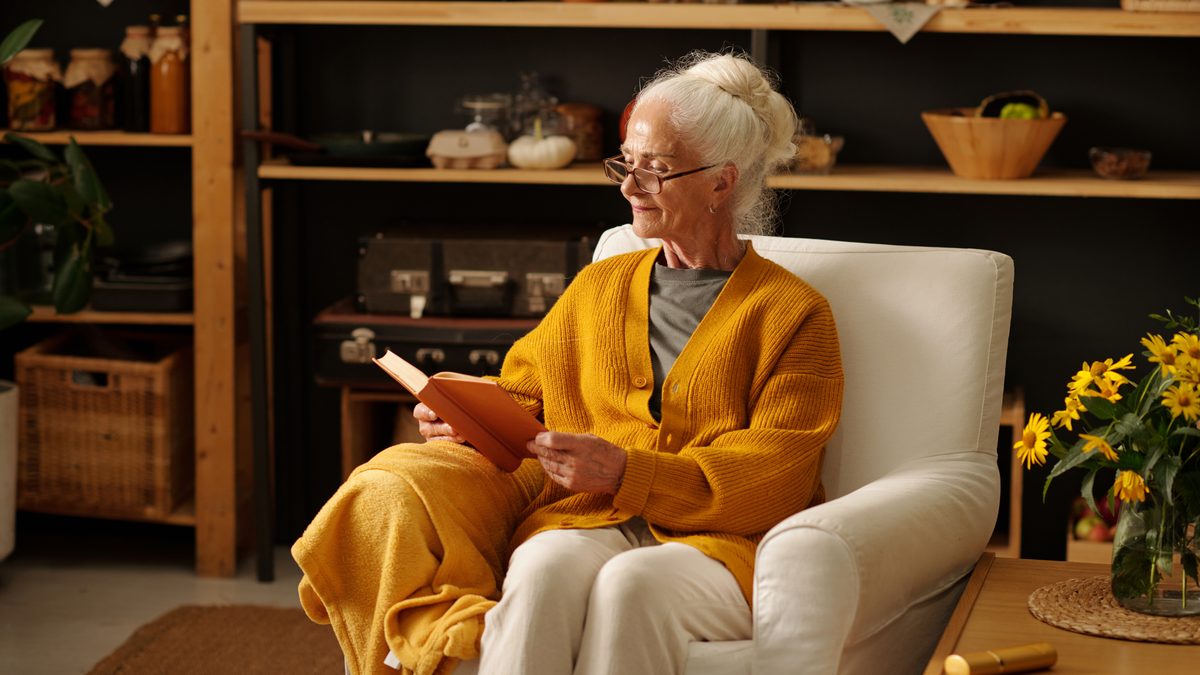 Aeltere Frau mit Brille und gelber Strickjacke sitzt mit einem Buch in einem hellen Sessel