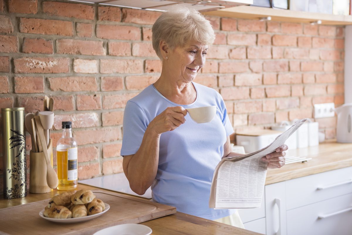 Aeltere Frau steht morgens in der Kueche, trinkt Kaffee und liest Zeitung