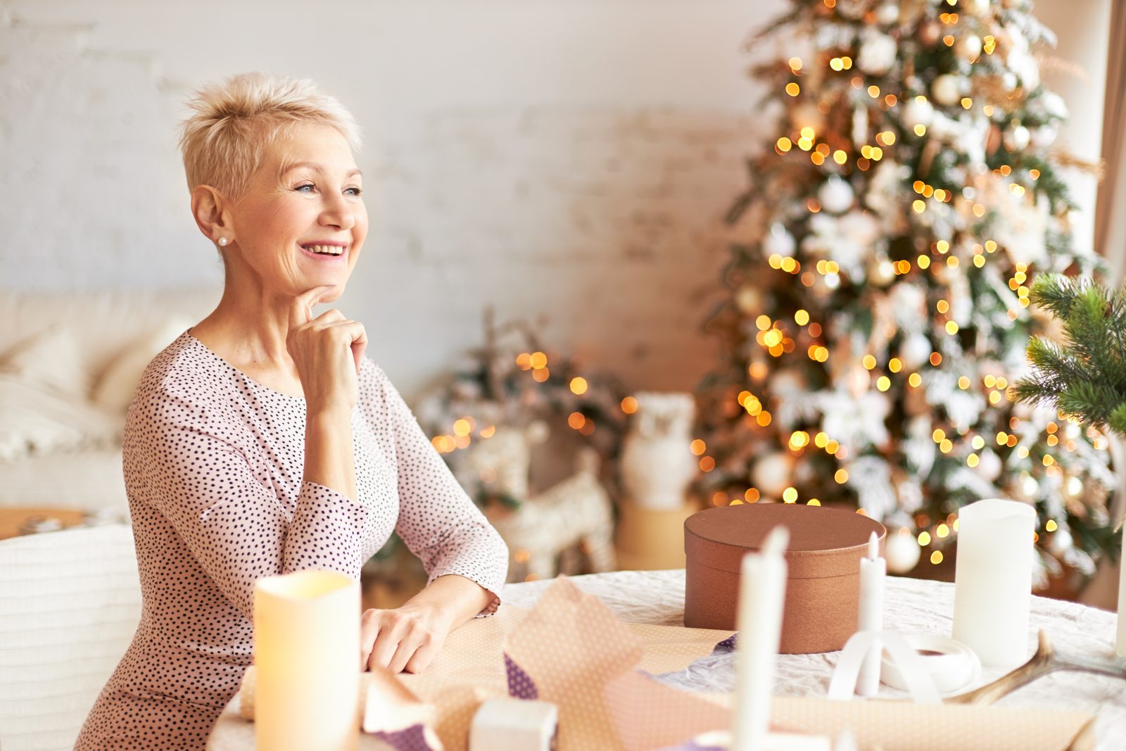 Aeltere Frau sitzt am Tisch und packt Geschenke in Packpapier ein, im Hintergrund ein geschmueckter Weihnachtsbaum