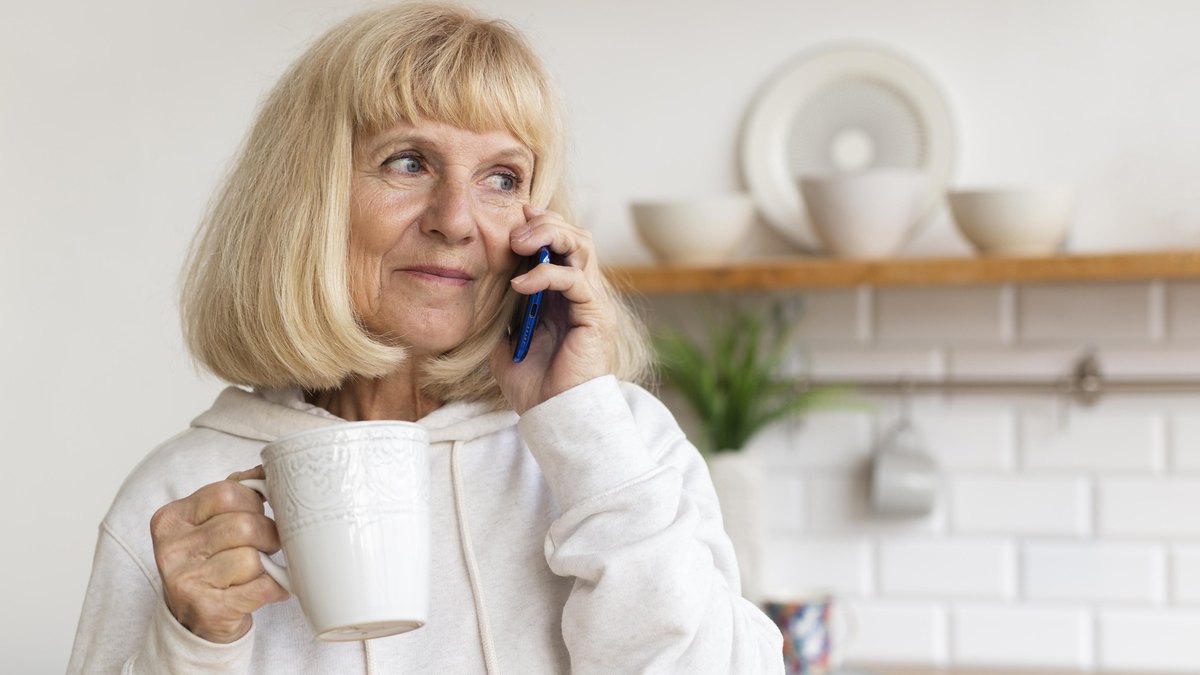 Aeltere Frau mit Kaffeebecher telefoniert in ihrer Kueche