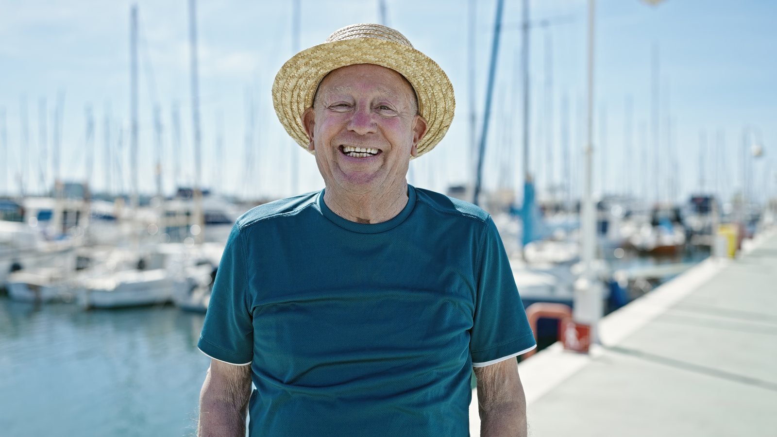 Aelterer Mann mit Strohhut steht laechelnd an einem sonnigen Tag am Hafen