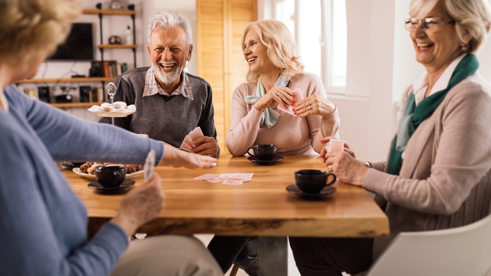 Vier aeltere Menschen sitzen lachend bei Kaffee und Gebaeck am Holztisch und spielen Karten