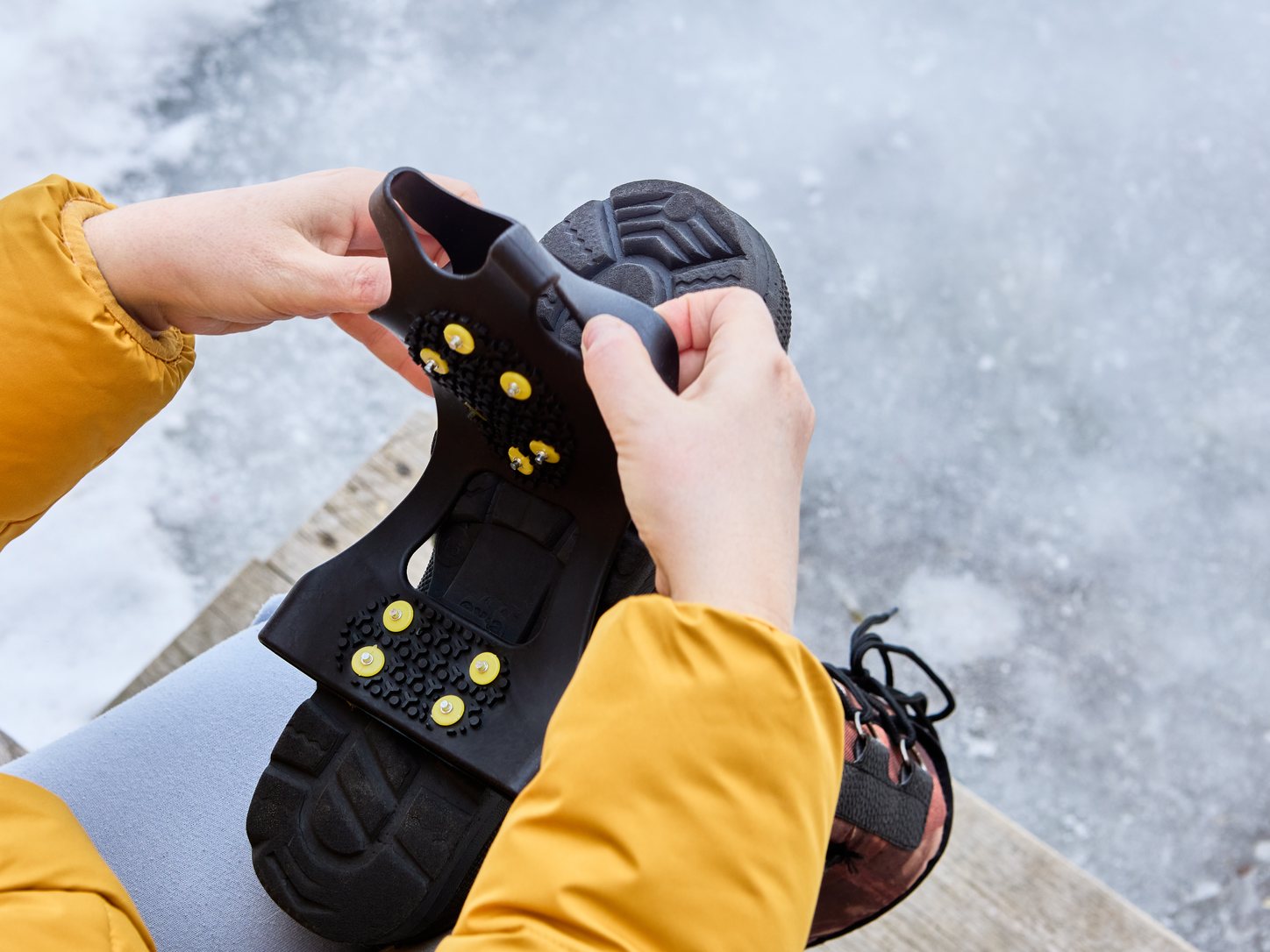 Person zieht im Sitzen einen Gummi-Ueberzieher mit Spikes ueber die Sohle eines Winterschuhs