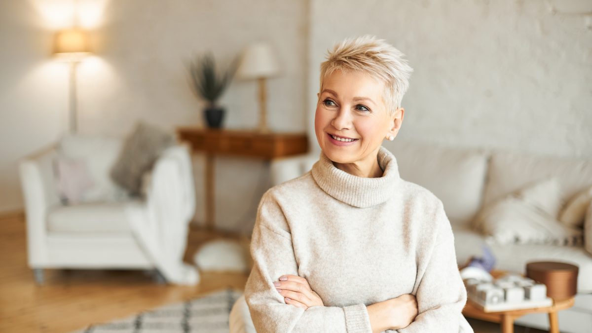 Aeltere Frau mit kurzen grauen Haaren in einem hellen Rollkragenpullover steht laechelnd im Wohnzimmer