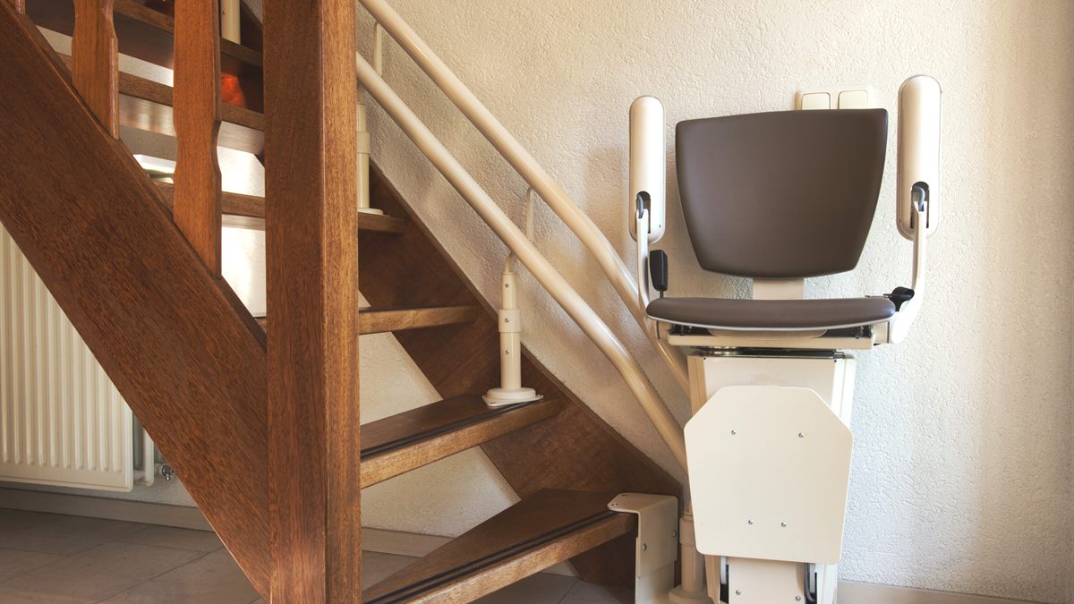 Sitzlift mit grauem Polstersitz und hochgeklappter Fussstuetze am unteren Ende einer Holztreppe im Wohnhaus, die Fahrschiene laeuft an der Wand entlang