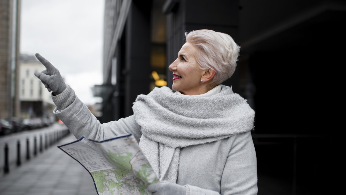 Aeltere Frau mit grauem Schal haelt einen Stadtplan und zeigt auf dem Gehweg in eine Richtung