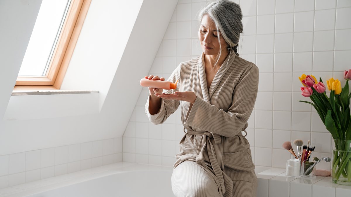 Seniorin im Bademantel sitzt im Bad auf dem Wannenrand und gibt sich Creme auf die Hand