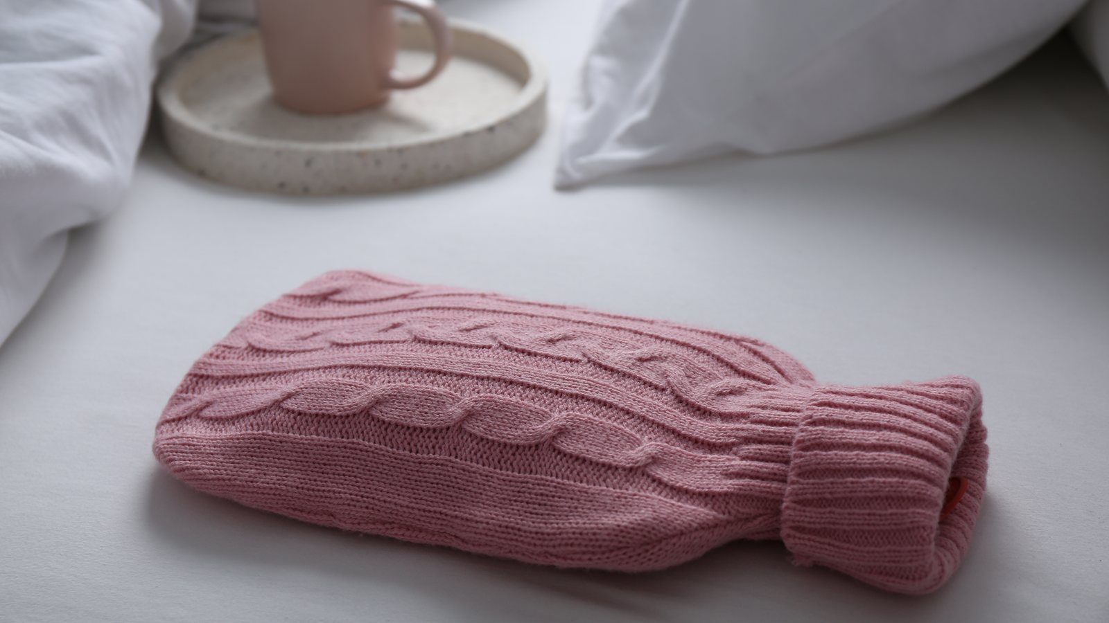 Waermflasche in einem rosa Strickbezug liegt auf dem Bett, dahinter eine Tasse Tee