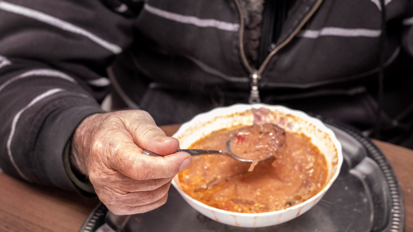Aeltere Hand fuehrt einen Loeffel ueber einer Schuessel mit warmer Suppe