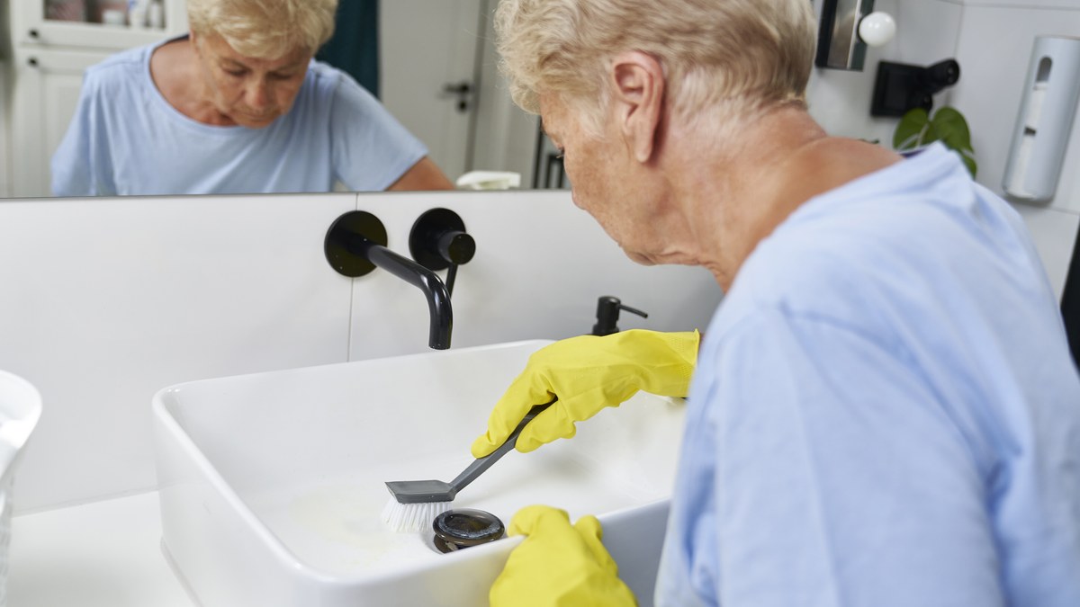 Aeltere Frau mit gelben Handschuhen reinigt das Waschbecken mit einer Buerste