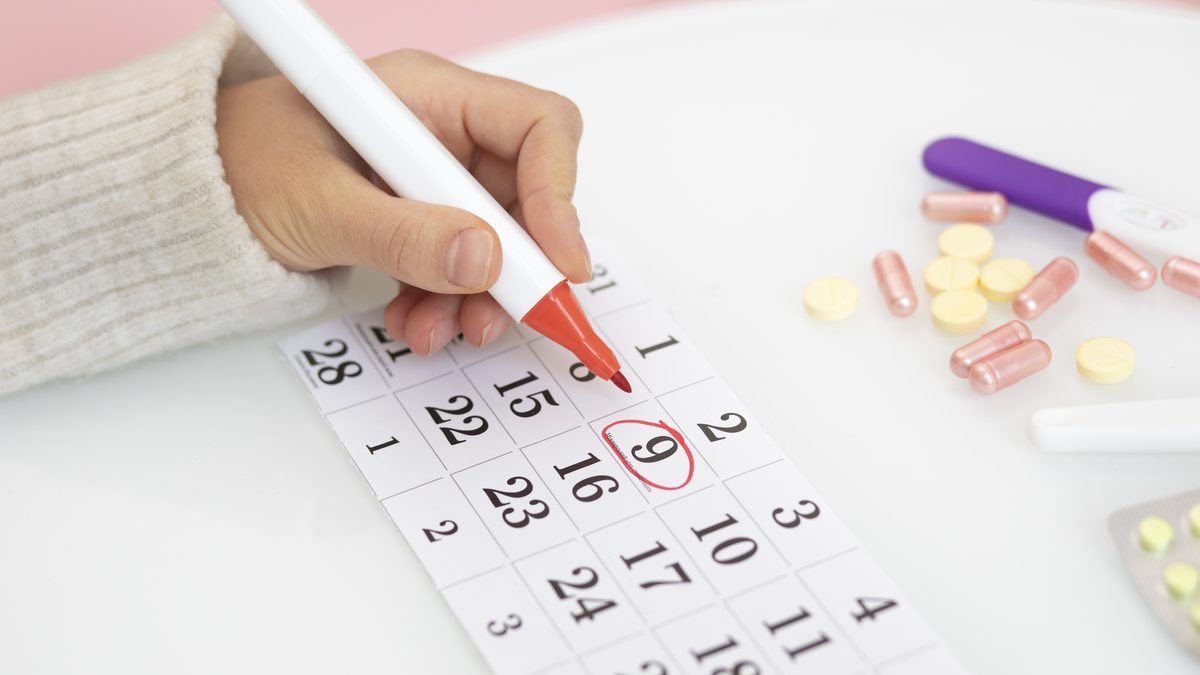 Hand markiert mit rotem Stift einen Tag im Kalender, daneben liegen Tabletten und Kapseln